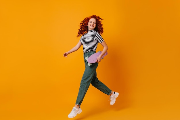 Aktives blauäugiges rothaariges Mädchen in weißen, stylischen Turnschuhen und Jeans, die auf orangefarbenem Hintergrund springen und lila Longboard halten