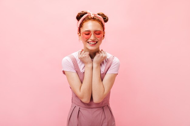 Aktives, fröhliches Mädchen mit rosa Brille und hellem Outfit lacht mit geschlossenen Augen auf rosa Hintergrund