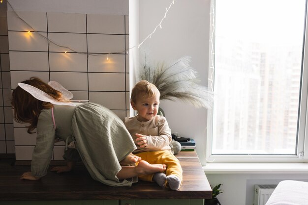 Aktives kaukasisches kleines rothaariges Mädchen im Kleid mit fairhaarigem Jungen klettert auf die Tischplatte in der Nähe des Fensters Kinderkonzept