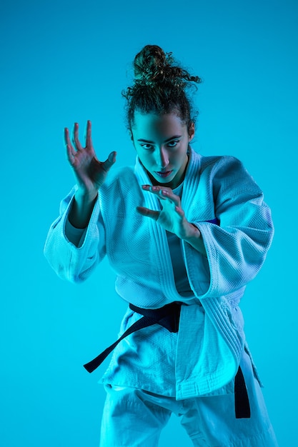Aktives Training. Professioneller weiblicher Judoist im weißen Judo-Kimono, der lokalisiert auf blauem neoniertem Studiohintergrund praktiziert und trainiert.