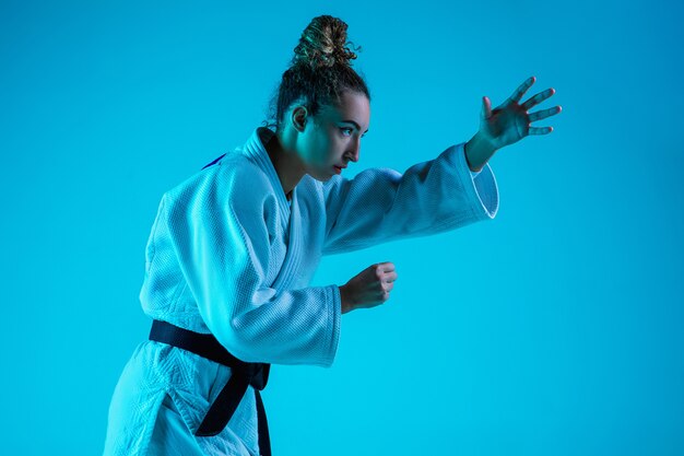 Aktives Training. Professioneller weiblicher Judoist im weißen Judo-Kimono, der lokalisiert auf blauem neoniertem Studiohintergrund praktiziert und trainiert.