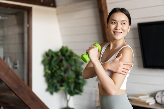 Aktives und gesundes asiatisches Mädchen mit fittem Körper, das zu Hause Fitnessübungen macht, Hanteln heben und...