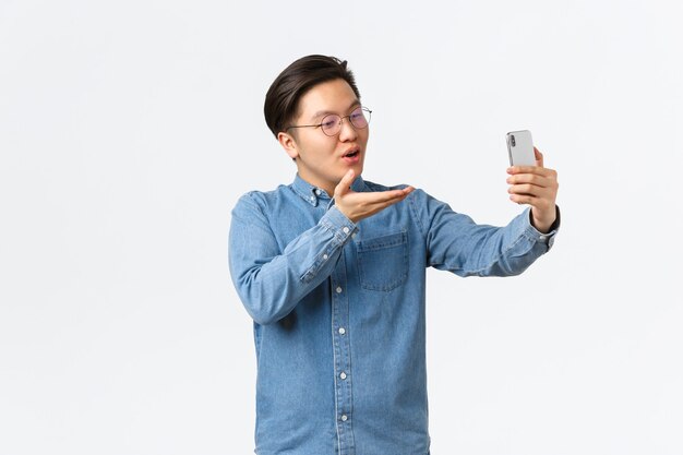 Alberner und süßer asiatischer Kerl, der lächelt, mit seiner Freundin über Smartphone, Videoanrufe oder Selfie spricht, Luftkuss an die Handykamera sendet, weißer Hintergrund romantisch steht.