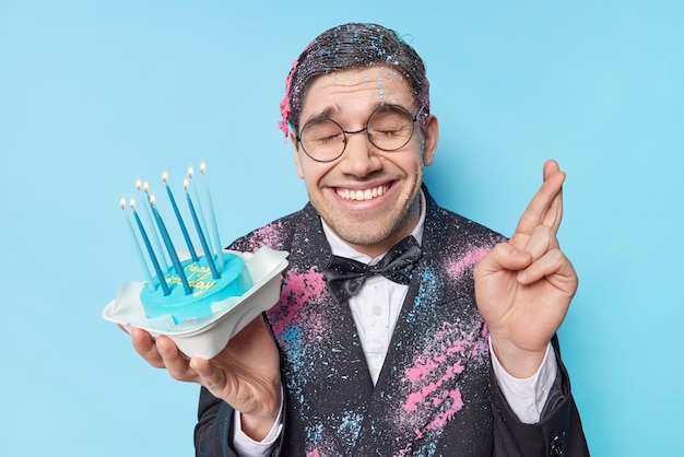 Alles Gute zum Geburtstag gutaussehender Mann kreuzt die Finger macht Wunsch hält Kuchen mit brennenden Kerzen in festlicher Kleidung gekleidet lächelt breit isoliert über blauem Hintergrund Partyzeit und Feierkonzept