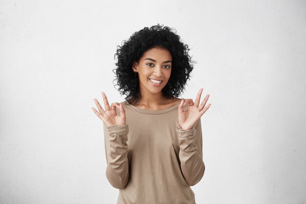 Alles ist perfekt. Glückliche positive dunkelhäutige Studentin, die mit beiden Händen eine gute Geste zeigt und gute Laune hat, nachdem sie alle Prüfungen am College erfolgreich bestanden hat.