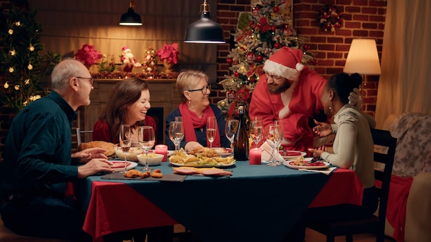 Als Weihnachtsmann verkleideter Mann spricht mit glücklicher Frau beim Weihnachtsessen zu Hause. Fröhliche afrikanisch-amerikanische Frau überrascht von Ehemann, der wie der Weihnachtsmann aussieht, während sie Winterurlaub feiert.