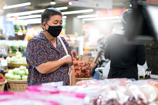 Alte asiatische Seniorin trägt eine Schutzmaske zur Verbreitung von Viren, während sie frisches Gemüse und Obst im Lebensmittelgeschäft im Supermarkt einkauft, neuer normaler Lebensstil, nachdem die Sperrung beendet ist