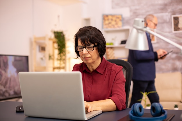 Alte Frau, die einen modernen Computer in ihrem Wohnzimmer benutzt, während ihr Mann im Hintergrund geht