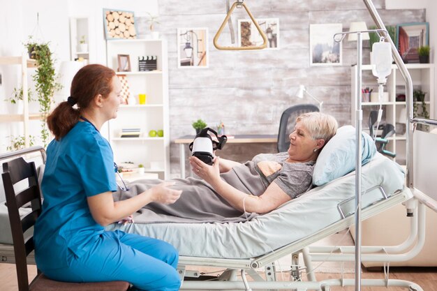 Alte Frau, die mit einer Virtual-Reality-Brille im Pflegeheim auf dem Bett liegt. Ärztin sitzt auf dem Bett.