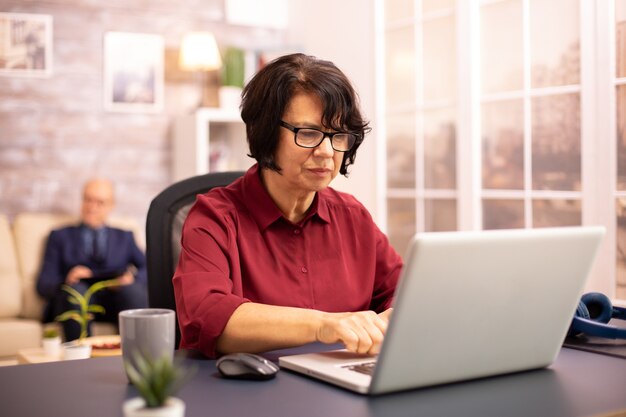 Alte Frau in ihren 60ern mit einem modernen Laptop in ihrem gemütlichen Haus spät am Abend