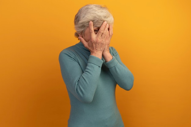 Kostenloses Foto alte frau mit blauem rollkragenpullover, der das gesicht mit den händen bedeckt, die durch die finger nach vorne schauen, isoliert auf der orangefarbenen wand mit kopierraum