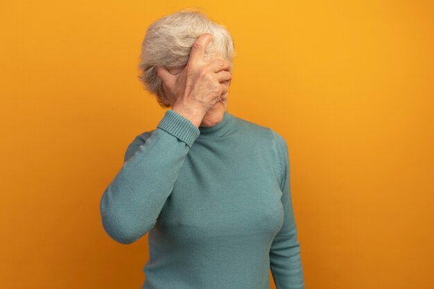Alte Frau mit blauem Rollkragenpullover, die Hand auf das Gesicht legt, isoliert auf oranger Wand mit Kopierraum