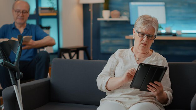 Alte Frau mit digitalem Tablet mit Touchscreen im Wohnzimmer. Ältere Person mit Krücken auf der Couch, die auf dem Gerät im Internet surft, um Spaß zu haben und den Ruhestand zu Hause zu genießen. Moderner Mensch