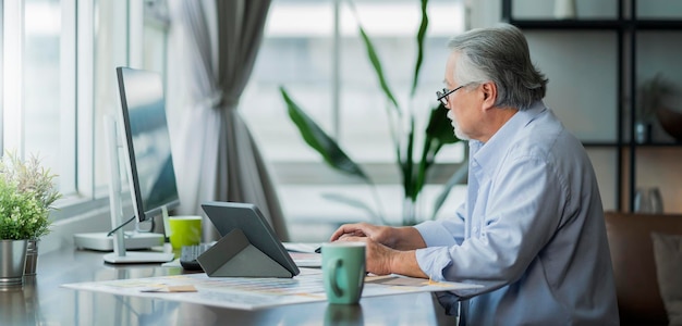 Alter asiatischer Senior im Ruhestand, der zu Hause arbeitet, hand ise Desktop- und Papierdokument am Arbeitstisch im Wohnzimmer, Innenhintergrund, asiatischer männlicher Senior, Arbeit zu Hause