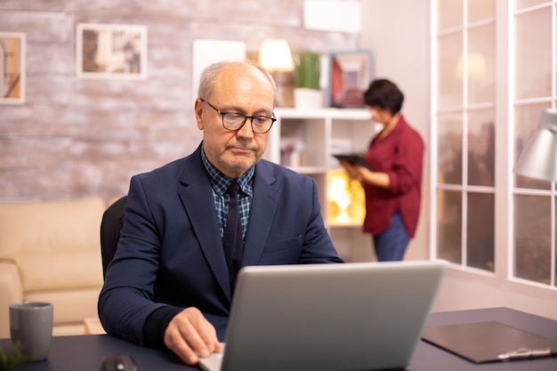 Alter mann in den 60ern, der im gemütlichen wohnzimmer an einem laptop arbeitet, während seine frau im hintergrund ist