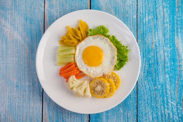 Amerikanisches Frühstück auf einem blauen Tisch mit Spiegelei, Salat, Kürbis, Gurke, Karotte, Mais, Blumenkohl und Tomate.