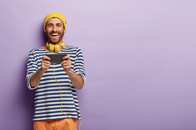 Amüsiert unterhaltener Typ spielt Videospiele auf dem Handy, in Freizeitkleidung, lächelt positiv, trägt Kopfhörer um den Hals
