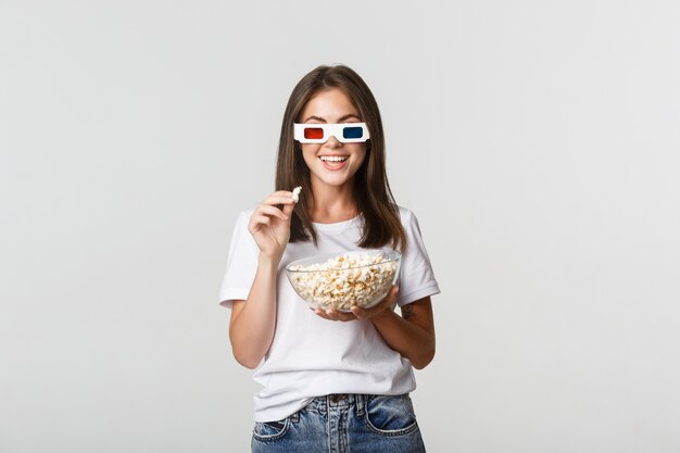 Amüsierte hübsche junge Frau in 3D-Brille, die Filme oder Fernsehserien sieht, Popcorn isst und begeistert lächelt.