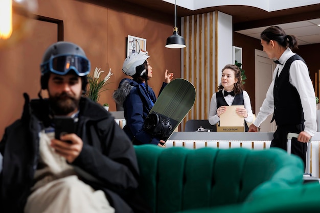 Kostenloses Foto an der rezeption helfen mitarbeiter einer jungen frau mit snowboardausrüstung bei der zimmerreservierung im winterresort. mann mit skihelm auf sofa benutzt sein mobiltelefon, während touristin eincheckt.