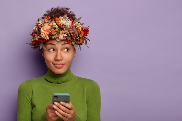 Angenehm aussehende nachdenkliche Herbstdame trägt natürlichen Kranz, benutzt Handy zum Online-Shopping, konzentriert beiseite, isoliert über lila Wand.