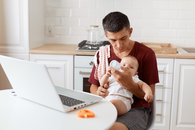 Angenehm aussehender dunkelhaariger, gutaussehender Mann, der ein lässiges T-Shirt mit einem Handtuch auf der Schulter trägt, mit Laptop am Tisch sitzt, ein Baby in den Händen hält und Babywasser zum Trinken gibt.