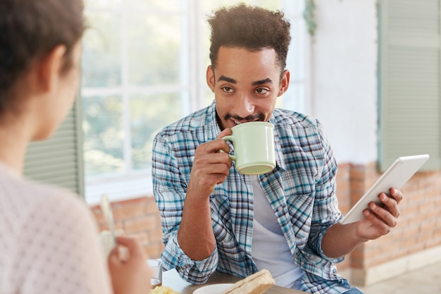 Angenehm aussehender Mann mit spezifischem Aussehen trinkt Kaffee mit Kuchen, spricht mit seiner Frau,