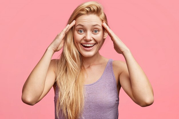 Angenehm aussehendes blondes blauäugiges junges weibliches Modell mit glücklichem Ausdruck, hält Hände auf Kopf, erinnert sich an einige positive Momente im Leben, gekleidet in lässigem T-Shirt.