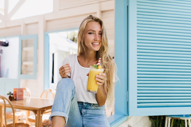 Angenehme blondhaarige Frau, die im Café mit Glas Saft sitzt. Erstaunliche gebräunte Dame im weißen T-Shirt lachend, während sie mit Cocktail aufwirft.