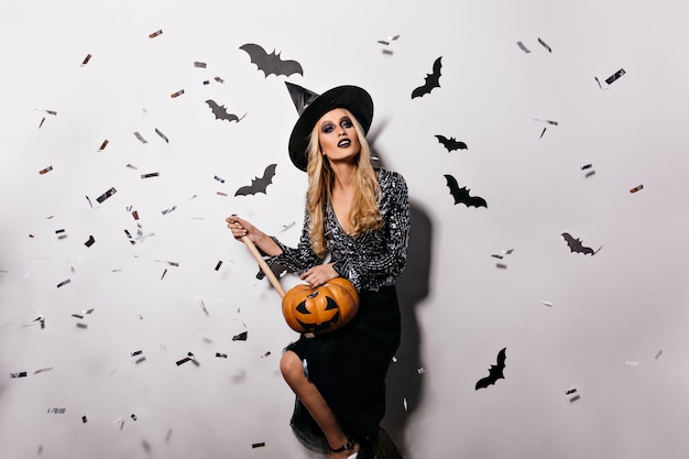 Angenehme junge Hexe, die mit Fledermäusen an der Wand aufwirft. Hübsches blondes Vampirmädchen, das Halloween-Kürbis hält.