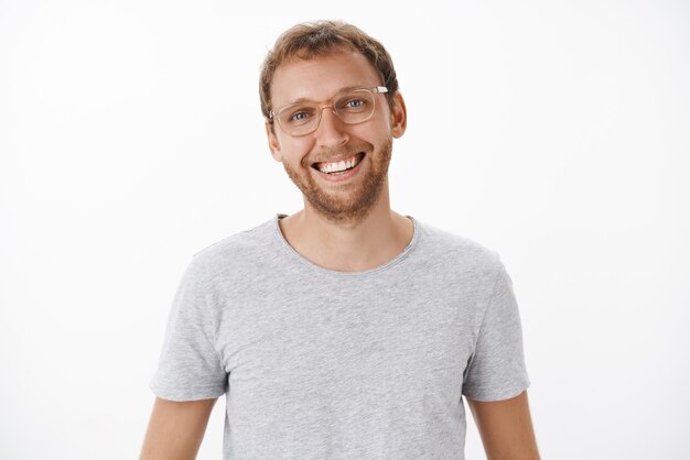 Angenehmer hübscher und freundlicher kaukasischer blonder Mann mit Borsten in der Brille und grauem lässigem T-Shirt, das freudig mit zufriedenem sorglosem Blick lächelt und Freizeit mit Familie genießt