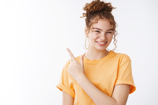 Angenehmes, hilfsbereites, gut aussehendes, freundliches Mädchen, das unordentliche lockige Haarknoten trägt, trägt ein orangefarbenes T-Shirt, das auf die obere linke Ecke zeigt, lächelt