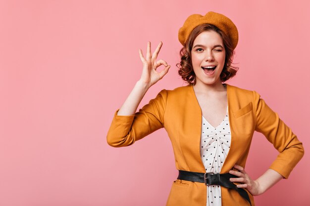 Angenehmes kaukasisches Mädchen, das okay Zeichen zeigt. Vorderansicht der schönen französischen Dame, die mit Hand auf Hüfte lokalisiert auf rosa Hintergrund aufwirft.