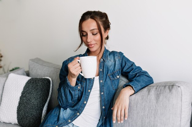 Angenehmes, stilvolles Mädchen mit gebräunter Haut und dunklem, gesammeltem Haar trägt Jeanshemd, hält Kaffee und ruht sich zu Hause aus