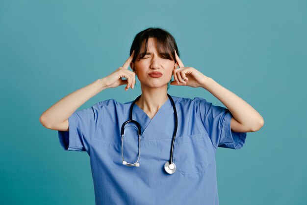 Angespannter Finger auf Kopf junge Ärztin mit einheitlichem Fith-Stethoskop isoliert auf blauem Hintergrund