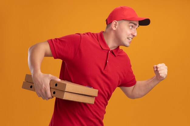 Angespannter junger Lieferbote, der Uniform mit Kappe hält, die Pizzakästen hält, die Laufgeste lokalisiert auf orange Wand zeigt