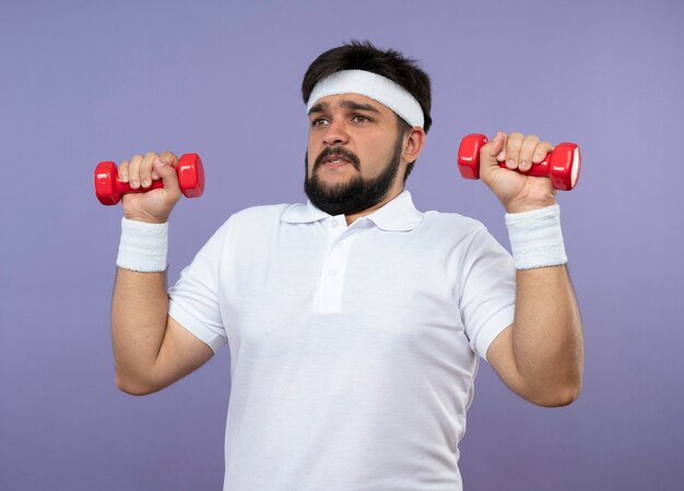 Angespannter junger sportlicher Mann, der Stirnband und Armband trägt, die mit Hanteln trainieren