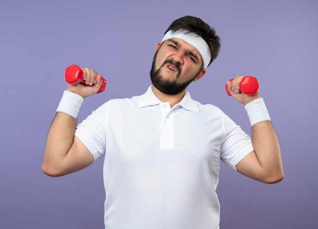 Angespannter junger sportlicher Mann, der Stirnband und Armband trägt, die mit Hanteln trainieren