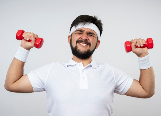 Angespannter junger sportlicher Mann, der Stirnband und Armband trägt, die mit mit Hantel, die auf weißer Wand lokalisiert wird, trainieren