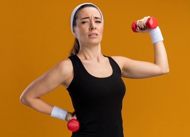 Angespanntes junges, hübsches, sportliches Mädchen mit Stirnband und Armbändern, das Hanteln hält und anhebt, die Hand auf der Taille isoliert auf oranger Wand halten?