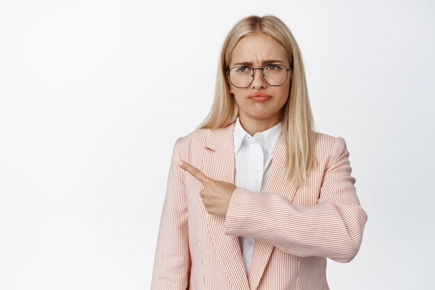 Angewiderte Geschäftsfrau in Brille und Anzug, die mit dem Finger nach links zeigt und schlechte Kriechdinge zeigt, die vor weißem Hintergrund stehen