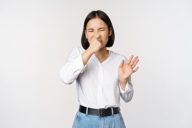 Angewidertes asiatisches Mädchen schließt die Nase und zeigt eine Ablehnungsgeste, die den schlechten Geruch verdirbt, der über weißem Hintergrund steht