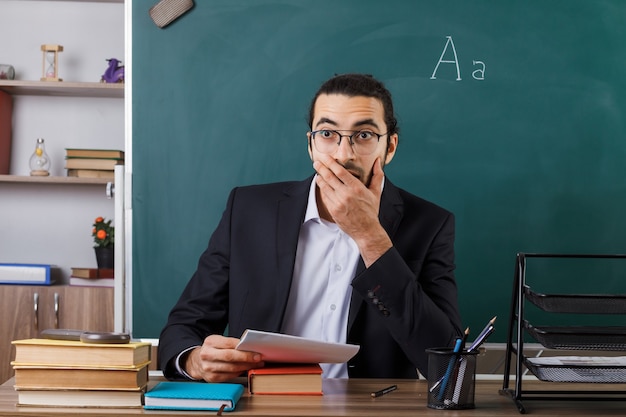 Angst bedeckter Mund mit Hand männlicher Lehrer mit Brille am Tisch sitzend mit Schulwerkzeugen im Klassenzimmer