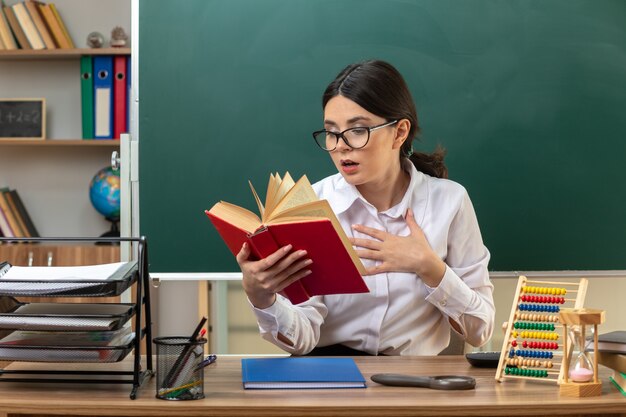 Angst, die Hand auf das Herz zu legen junge Lehrerin, die eine Brille trägt und ein Buch liest, das am Tisch mit Schulwerkzeugen im Klassenzimmer sitzt?