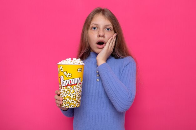 Angst, die Hand auf die Wange zu legen, schönes kleines Mädchen, das einen blauen Pullover trägt, der Popcorn-Eimer hält?