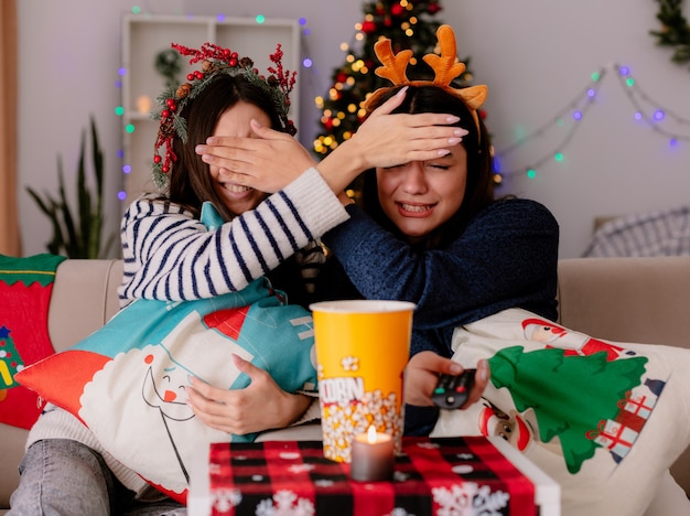 Angst hübsche junge Mädchen mit Stechpalmenkranz und Rentier Stirnband schließen sich die Augen mit der Hand auf Sesseln Weihnachtszeit zu Hause sitzen