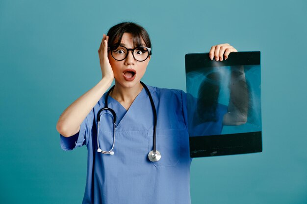 Angst packte den Kopf mit Röntgenstrahl Junge Ärztin mit einheitlichem Fith-Stethoskop isoliert auf blauem Hintergrund