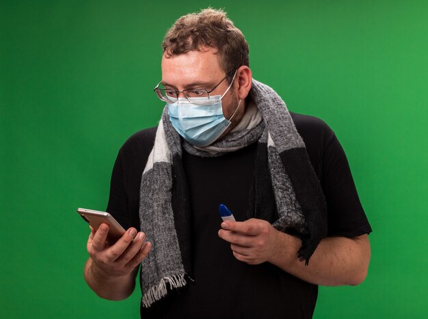 Angst vor einem kranken Mann mittleren Alters, der eine medizinische Maske und einen Schal trägt, der ein Thermometer hält und das Telefon in seiner Hand isoliert auf grüner Wand betrachtet