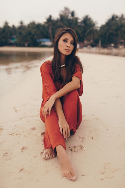 Anmutige Frau, die am Strand aufwirft, sitzt auf Sand im roten Kleid