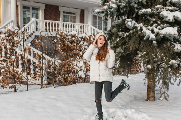 Anmutige Frau in zerrissenen Jeans, die am Wintertag auf der verschneiten Straße tanzen. Außenporträt der raffinierten europäischen Frau in der weißen Jacke, die im Hof neben Fichte herumalbert.