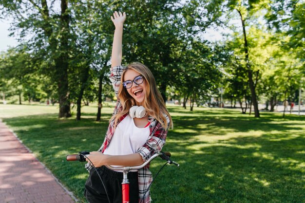 Anmutiges blondes Mädchen, das Aufregung ausdrückt. Außenfoto der fröhlichen weißen Dame mit dem Fahrrad, das auf Park aufwirft.